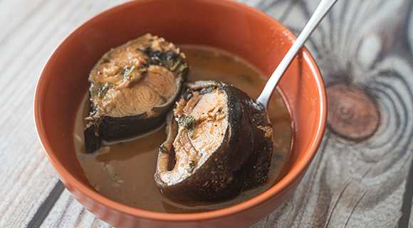 fish pepper soup in a orange ceramic bowl with a metal spoon on a natural wooden table