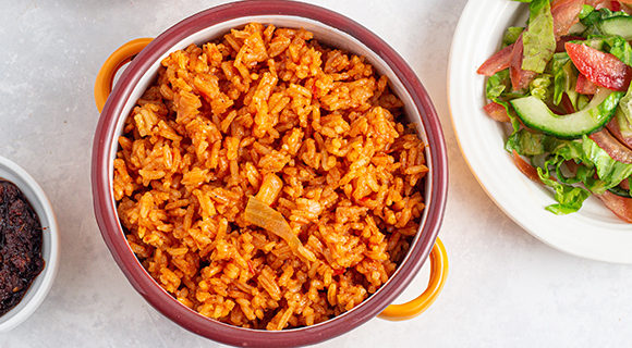 a red ceramic bowl of jollof rice with other white plates of salad next to it 